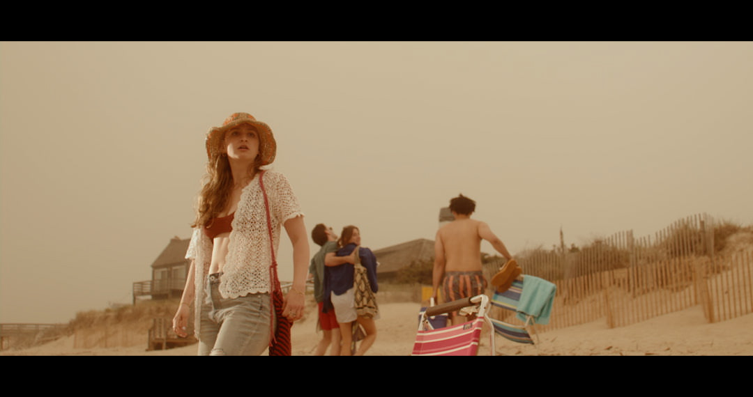 A group of people are on a beach. In the background we see a man lugging some collapsible beach chairs and a couple walking together away from shore. In the foreground of the image is a woman staring at the sea with what appears to be concern.